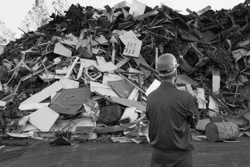 A man wearing a safety helmet and dark work clothes stands with his back to the camera, observing a large pile of mixed waste materials, including broken furniture, electronic appliances, and construction debris, situated outdoors on a paved surface. The debris appears to consist of various materials such as plastic, metal, and wood, with some items stacked haphazardly and others partially collapsed. In the background, sparse trees and an overcast sky are visible, suggesting a rural or industrial setting. The scene reflects an environment where tidying or clearance work is being carried out, likely in connection with rubbish removal services offered by Crystal Palace House Clearance in the local area of South London, including nearby postcode regions. The overall tone underscores a professional approach to waste disposal, with the focus on an outdoor site awaiting clearance or collection.