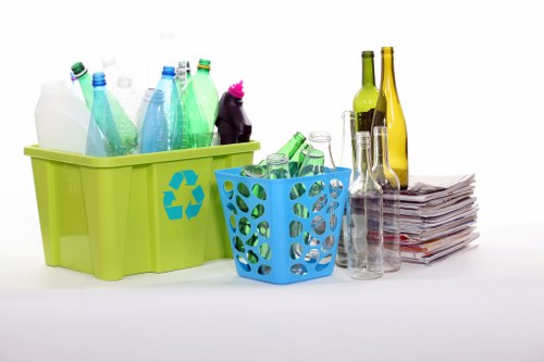 A collection of clear glass and plastic bottles, some with green and yellow tints, are arranged alongside a stack of newspapers or magazines, all placed on a plain white surface. In the foreground, there is a larger lime-green plastic bin with a blue recycling symbol on its side, containing several empty, transparent and coloured bottles, some with labels removed. To the right of the green bin, a smaller blue plastic basket with circular cut-out patterns holds more empty transparent bottles. The bottles vary in shape, with some having narrow necks and others wider openings, and the surface of the bottles shows some reflections, indicating a smooth, glass or plastic finish. Behind the bottles, a stack of folded newspapers or magazines rests on the white surface, with some slight crumpling visible. The background is entirely white and uncluttered, emphasizing the objects in the foreground, which suggest the context of rubbish or waste collection, possibly related to household recycling activities in the UK, such as those conducted by Crystal Palace House Clearance in the local SE19 area.