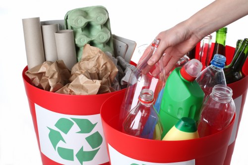 A pair of bright red recycling bins, each featuring a green recycling symbol, are filled with various waste materials. The left bin contains several cardboard tubes, crumpled brown paper, and a green egg carton, indicative of recyclable packaging and paper waste. The right bin holds an assortment of empty plastic bottles, including clear, green, and blue-tinted containers, alongside a yellow plastic bottle and a few dark-colored glass bottles, all arranged at different angles. A person's hand with a fair complexion is seen reaching into the right bin, grasping a clear plastic bottle. The scene appears to be set outdoors on a neutral surface, possibly a driveway or pavement, with no background details visible. The waste collection demonstrates typical household recyclable items, aligning with the services provided by Crystal Palace House Clearance, serving the Crystal Palace area within London postal districts.