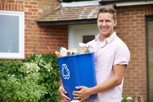 A young man with short light brown hair, wearing a light pink short-sleeved shirt, is standing outdoors in front of a brick residential building with a white window and a tiled roof. He is holding a large blue recycling bin with a white recycling symbol on the side, filled with various waste items including cardboard, paper, and plastic bottles. The bin is positioned slightly to his right, and he has a friendly, neutral facial expression. In the background, there are green bushes and flowering plants, indicating a garden or front yard setting in Crystal Palace, South London, consistent with local rubbish removal services provided by Crystal Palace House Clearance. The lighting suggests a bright, sunny day.