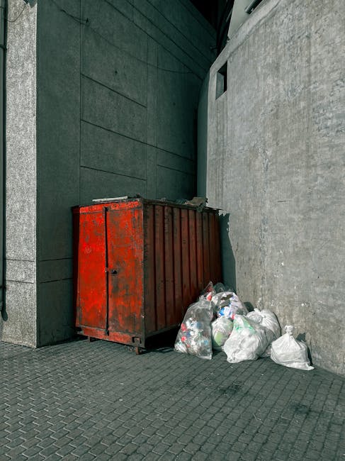 A collection of overflowing rubbish bins and scattered waste surrounding a large metal recycling container situated on a paved urban street. The recycling bin, marked with blue signage indicating it i