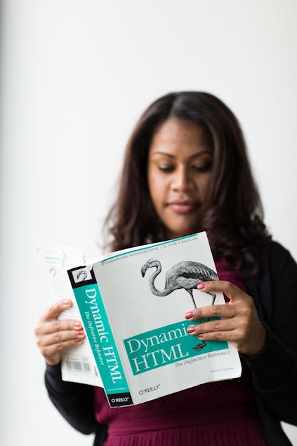 A woman with medium-length dark hair and light brown skin is holding a book titled 'Dynamic HTML' with a flamingo illustration on the cover. She is dressed in a dark blazer over a maroon top, standing
