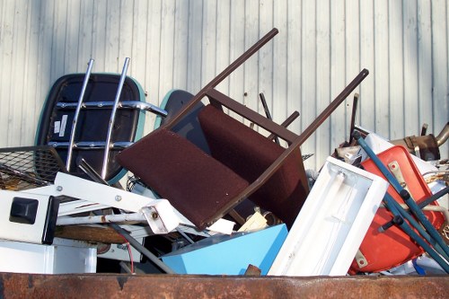 A large pile of assorted discarded household items and rubbish stacked together outdoors, featuring a blue plastic storage container, a white-framed window, a red plastic container, chairs with dark brown seats and metal frames, and a black plastic basket, all resting on an uneven surface possibly on a driveway or yard next to a corrugated metal wall, suggesting the scene of a clear-out by Crystal Palace House Clearance. The items appear used and discarded, forming a jumble of materials and textures typical of household waste or debris awaiting rubbish removal service in the Crystal Palace area. The lighting is natural, highlighting the variety of colours and surfaces, with some items overlapping each other in a haphazard arrangement consistent with clearing or disposal activities.